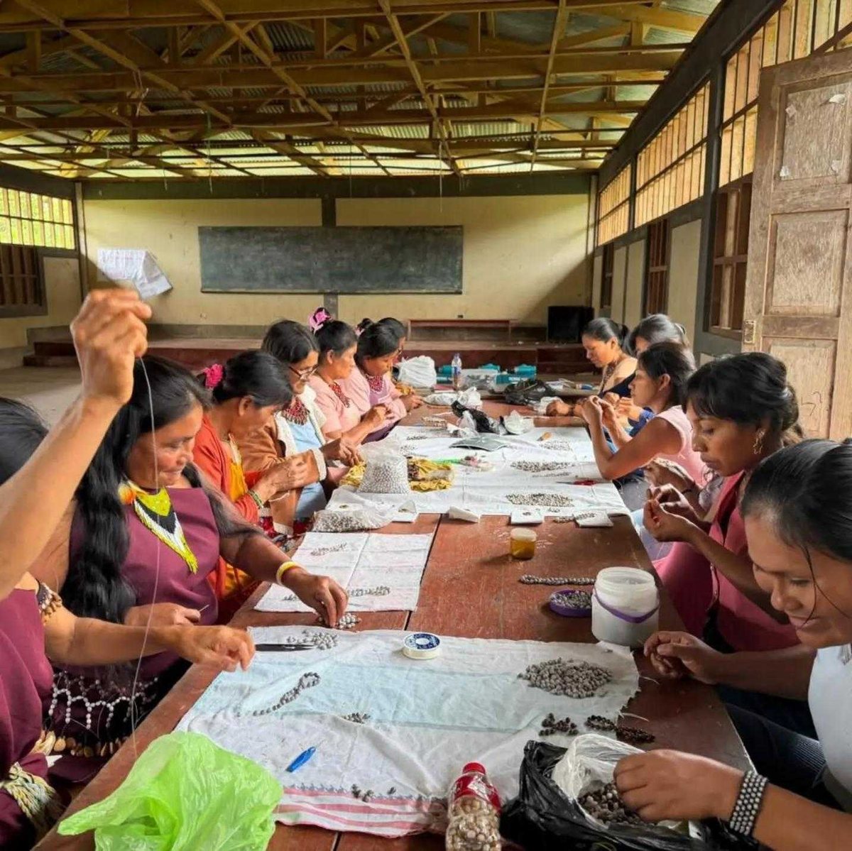 Mujeres awajún sentadas a una mesa larga enhebrando semillas; sobre la mesa hay montones de semillas, hilo y tijeras.